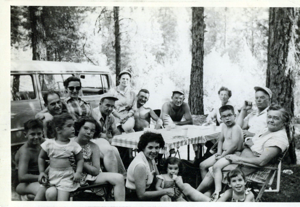 A large group of people posing for a black and white photograph at a picnic table in a wooded area near a station wagon.