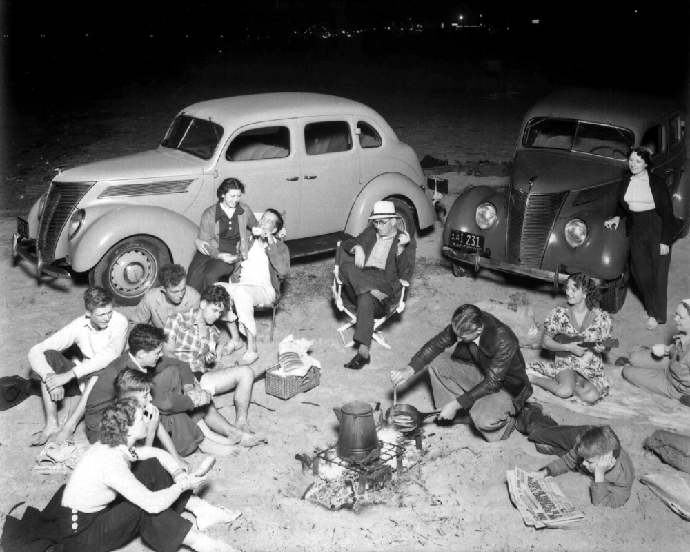 A group of people sitting on the beach around a small campfire with two parked vintage cars behind them.