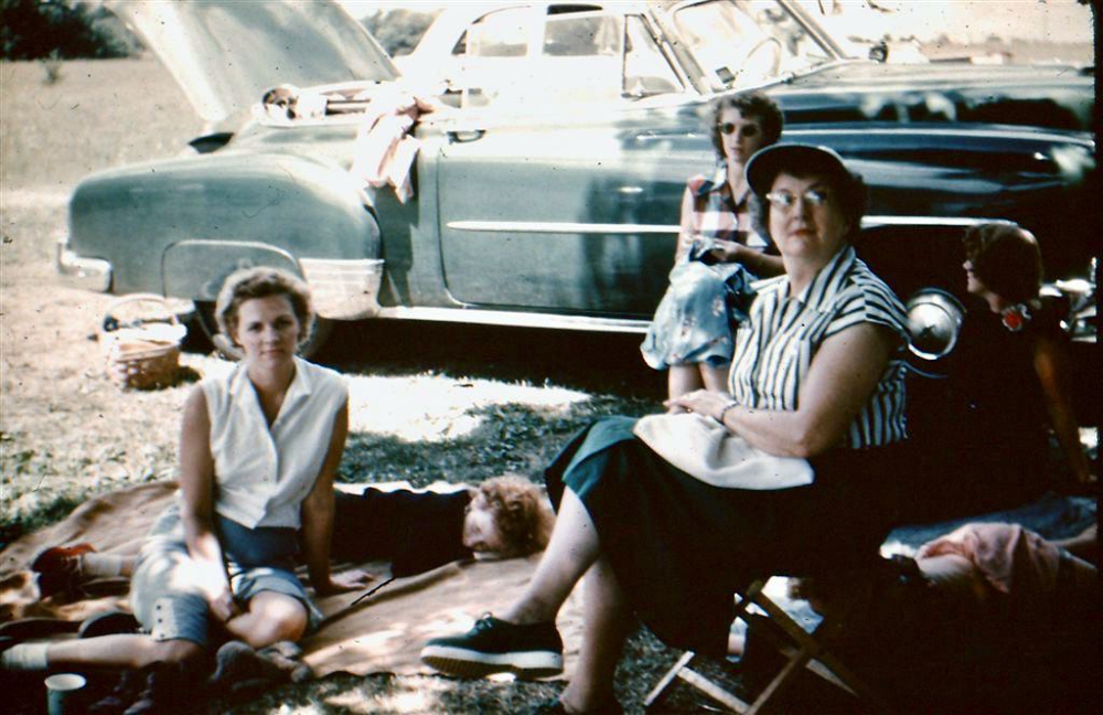 A group of people sitting on blankets and a folding chair in front of a vintage car parked in a field.