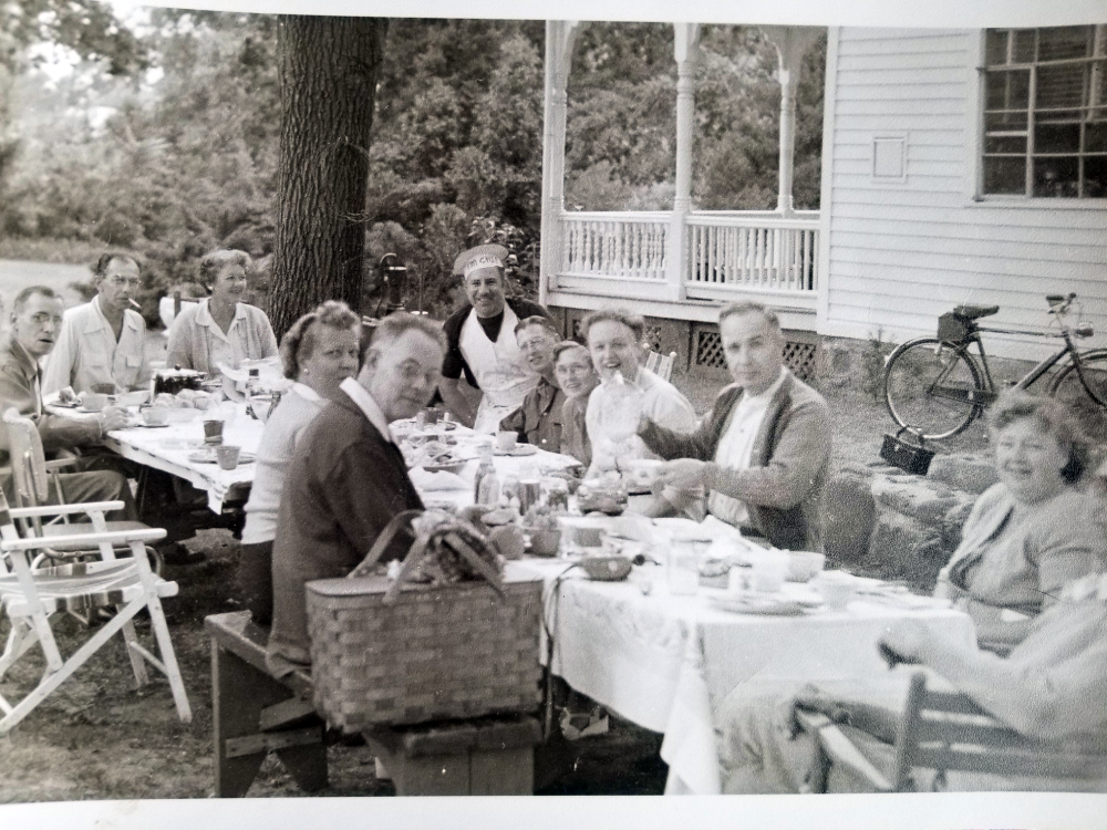 A group of people sitting at a long, outdoor table set for a meal near a house with a porch.