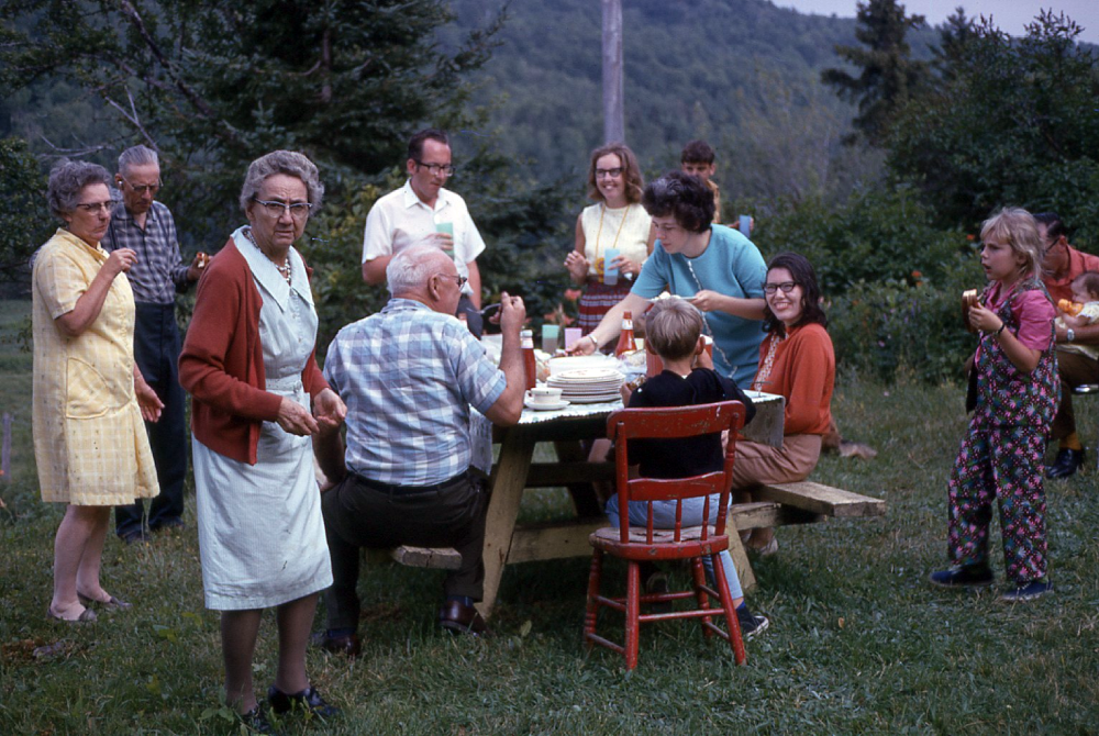 A group of people gathering for an outdoor meal at a picnic table in a grassy field.