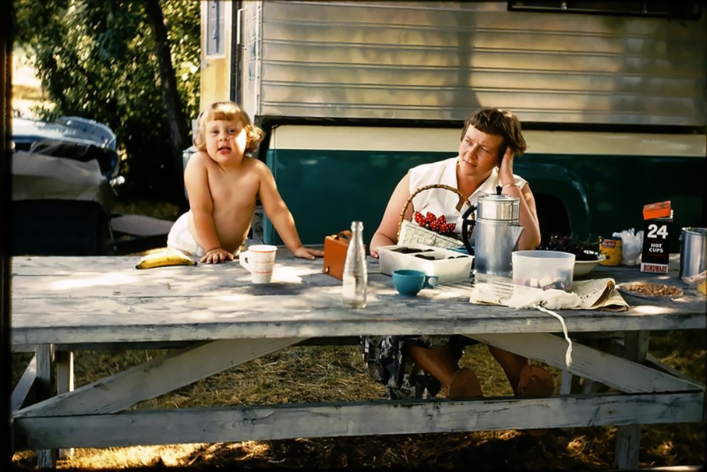 A woman and a young child sit at a picnic table outside a vintage camper trailer.