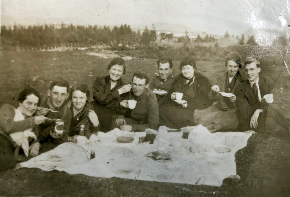 A black and white photograph of eight people picnicking on a blanket in a field.