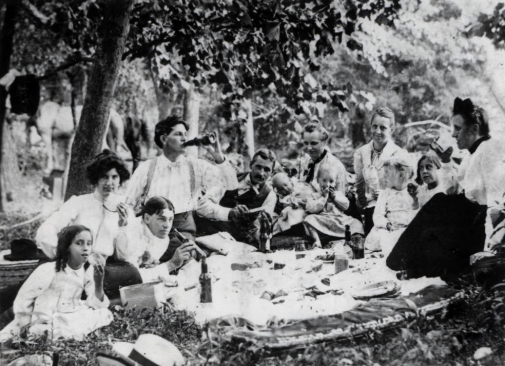 A large group of people having a picnic in a wooded area, with some drinking from bottles and others eating.