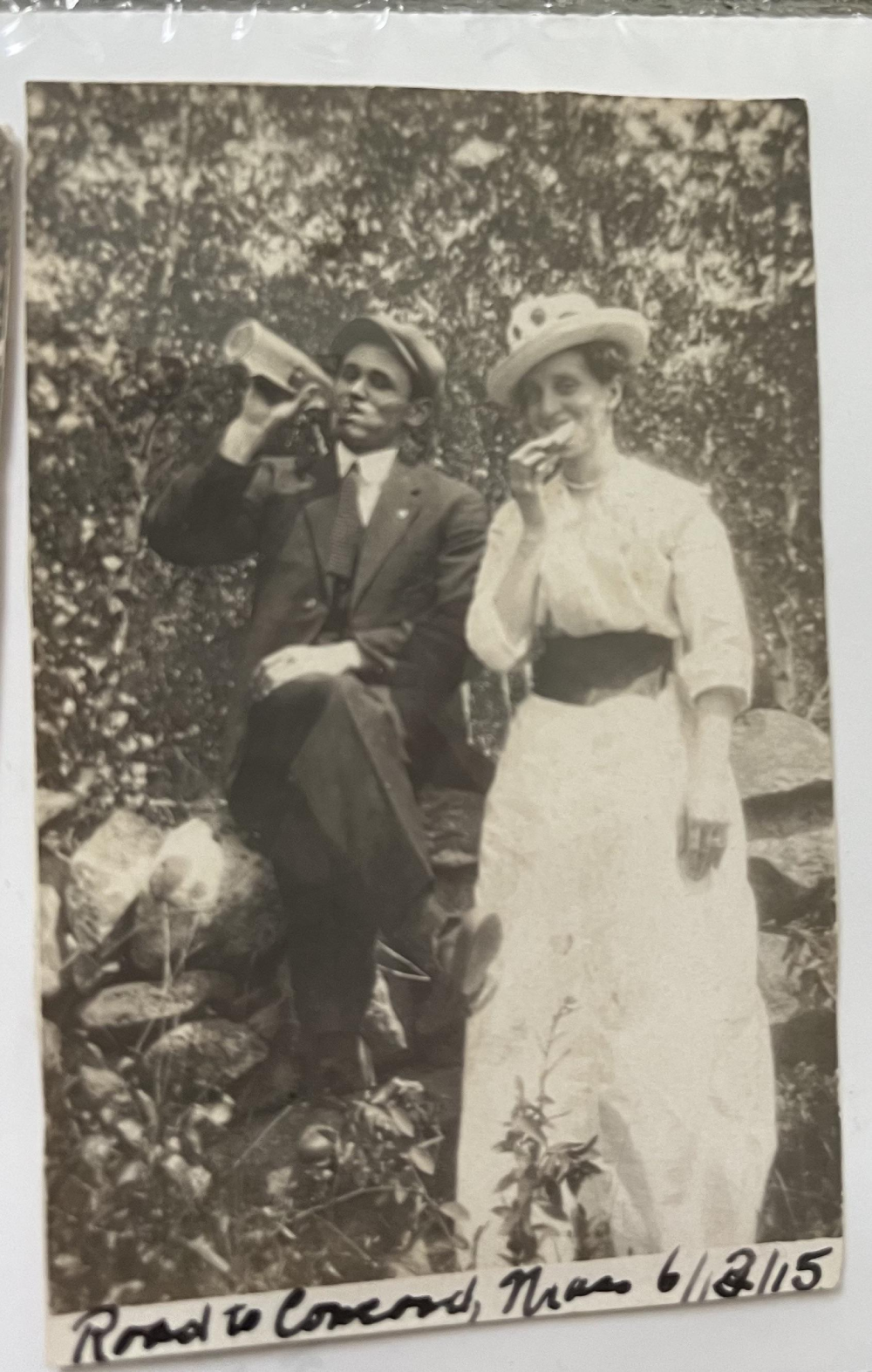 A black-and-white vintage photograph of a man in a suit drinking from a bottle and a woman in a long dress and hat eating, with the handwritten caption “Road to Concord, Mass 6/12/15” at the bottom.