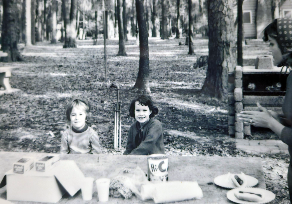Two children sit at a picnic table outdoors, with a woman standing nearby holding a plate.