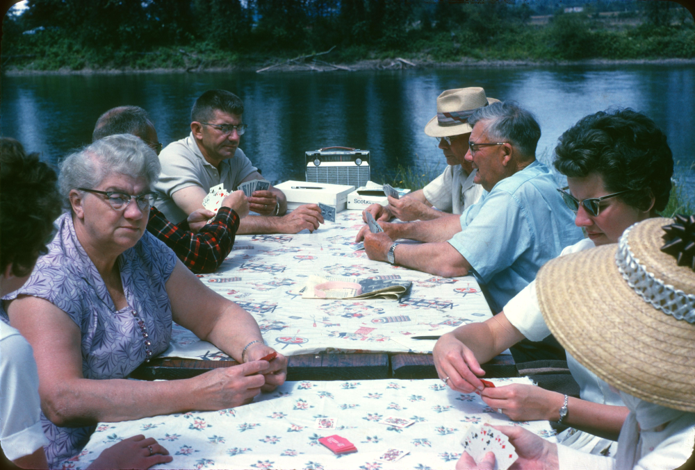 A group of six people sit around a picnic table playing cards outdoors near a river.