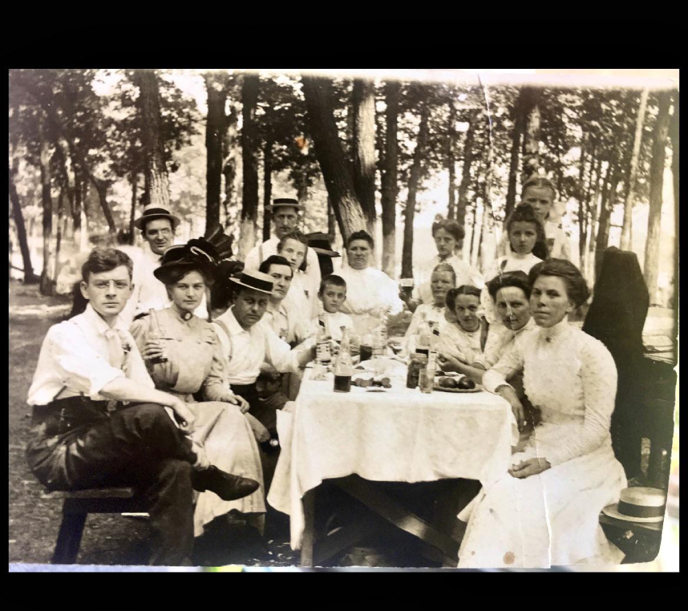 A large group of people in late 19th-century attire gathered around a table set for an outdoor meal in a wooded area.