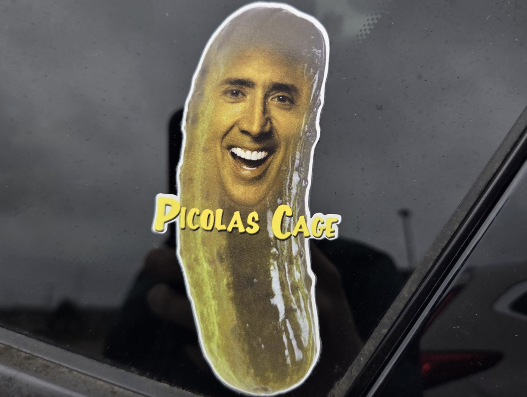 A sticker on a window depicting a pickle with the face of actor Nicolas Cage, captioned “Picolas Cage.”