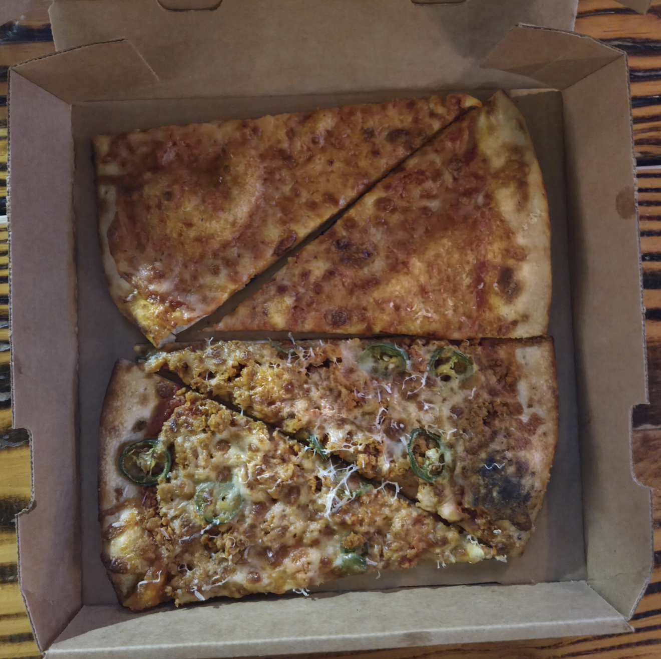 An open pizza box with two slices of cheese pizza and two slices of pizza topped with jalapeños and shredded cheese, placed on a wooden table.
