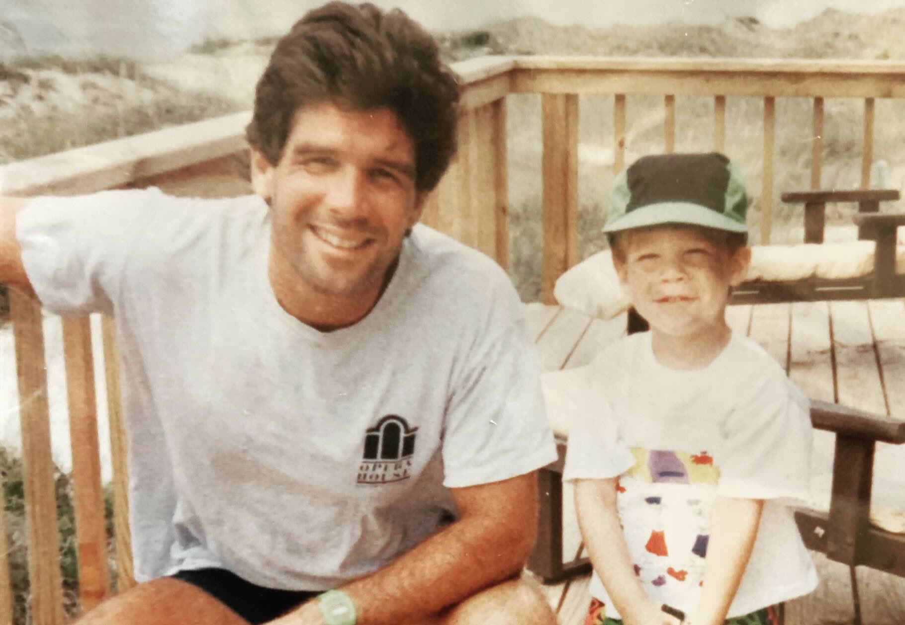A man and a young boy smiling together on a wooden deck.