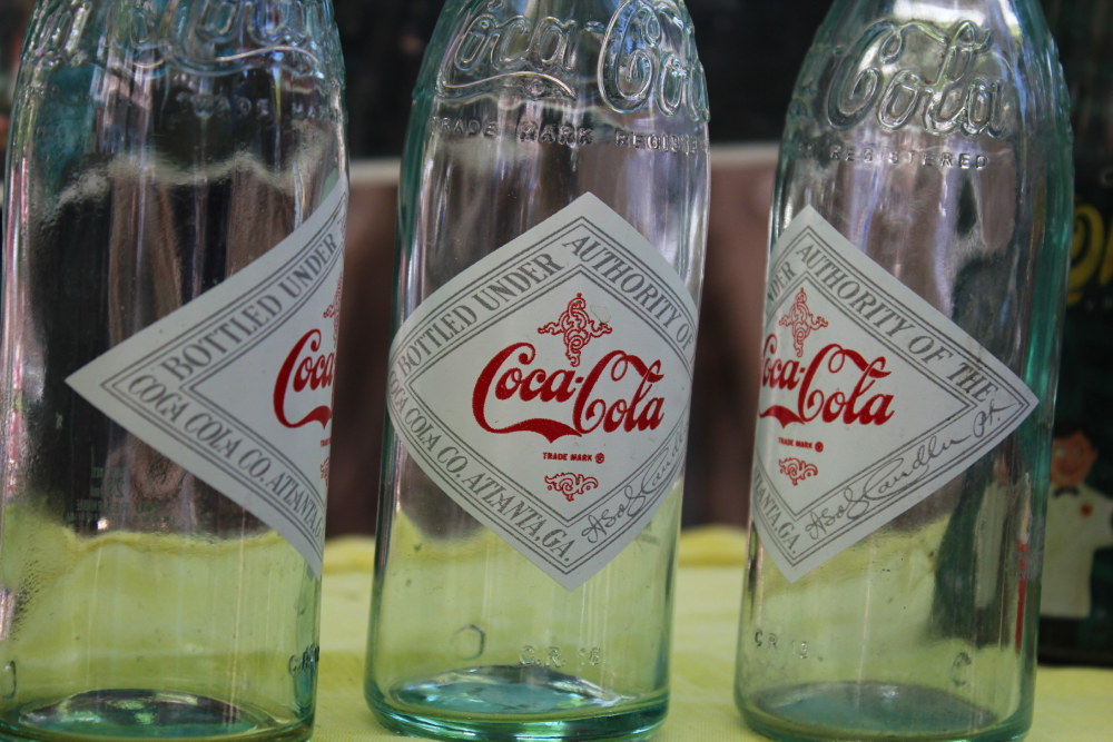 Three vintage clear glass Coca-Cola bottles with diamond-shaped white labels.