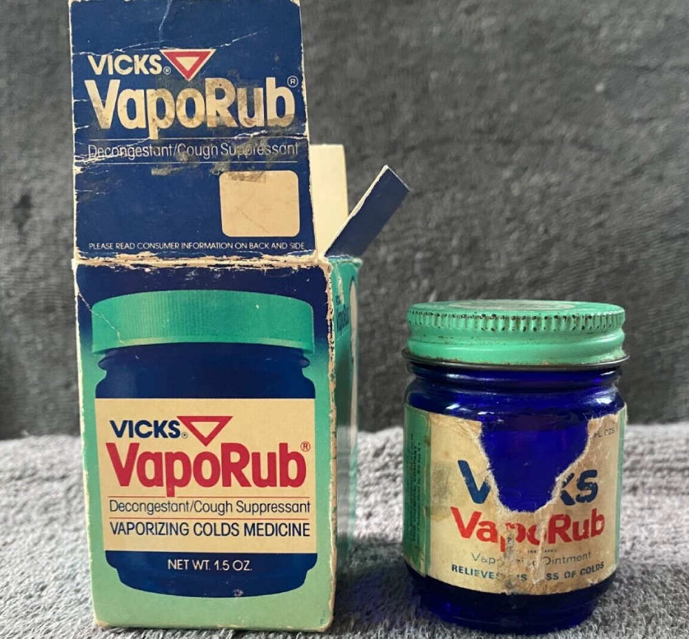 An open Vicks VapoRub cardboard box next to its matching blue glass jar with a distressed label.