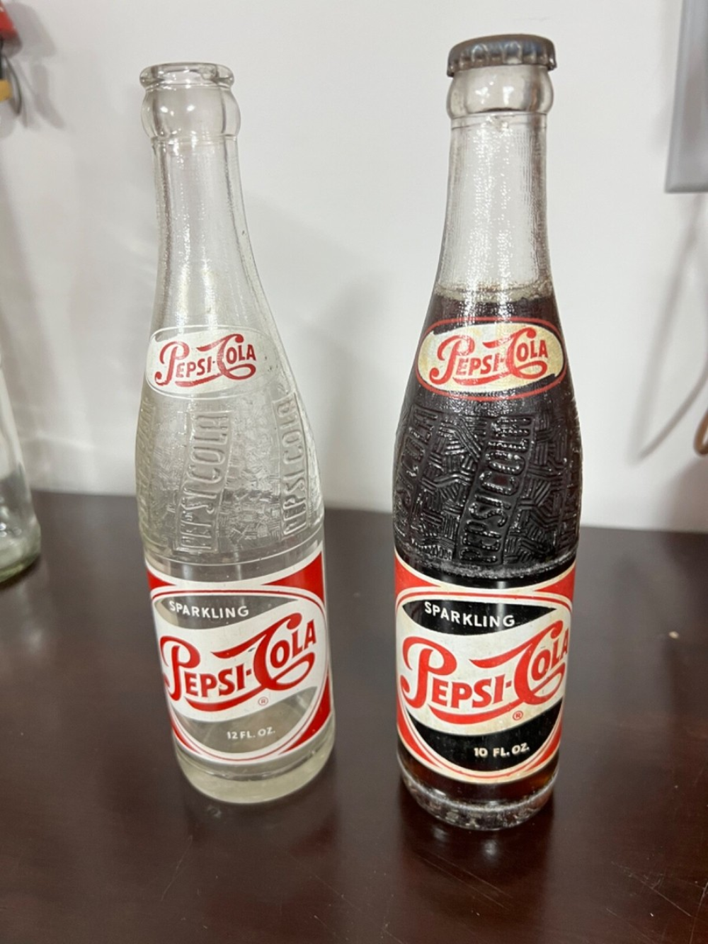 Two vintage Pepsi-Cola glass bottles, one empty and one full, standing side-by-side on a wooden surface.