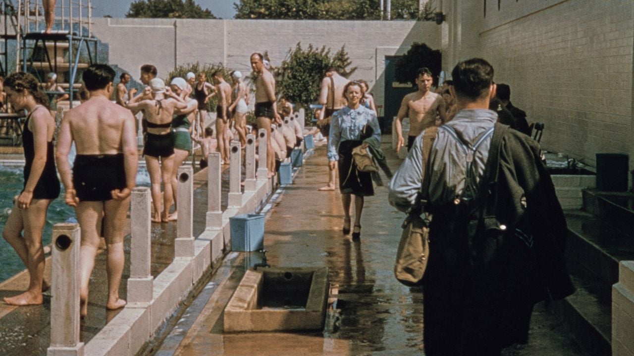 People in swimsuits gather by a busy outdoor pool, some standing and others walking. A woman in blue walks toward the camera, while a man carrying items walks away. The scene is lively and sunlit.