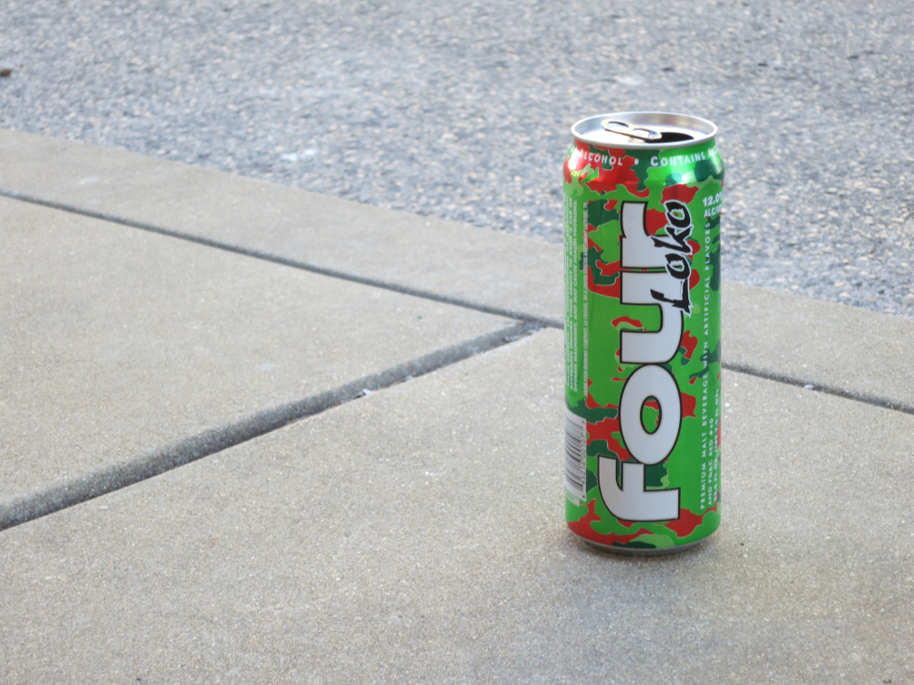 An open can of green and red camouflage-patterned Four Loko malt beverage sitting on a concrete sidewalk.
