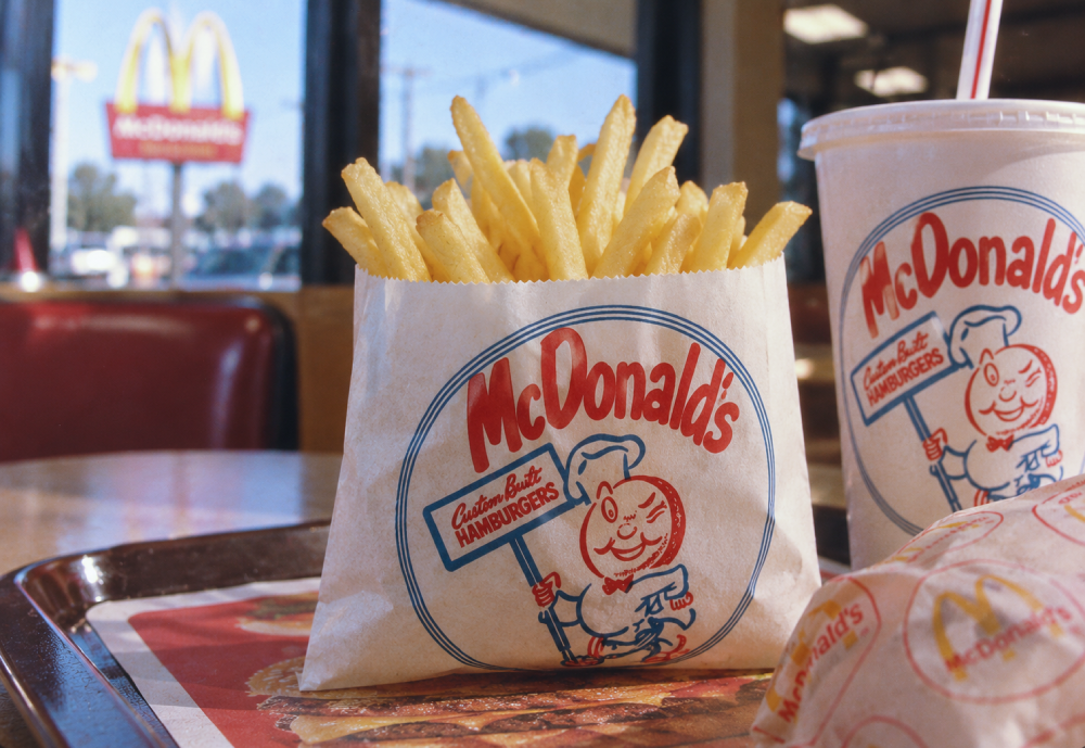 A paper bag filled with French fries, a paper cup with a straw, and a wrapped burger on a tray, with a McDonald’s sign visible through the restaurant window.