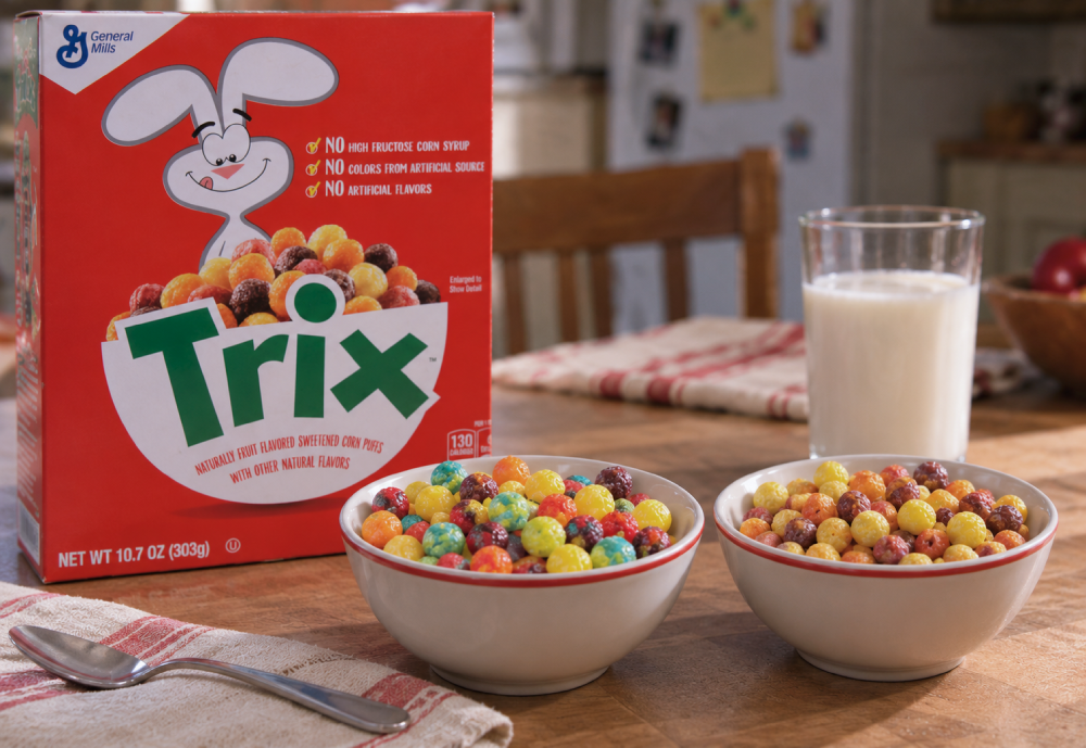 A box of Trix cereal sitting on a wooden table next to two bowls of colorful cereal puffs and a glass of milk.