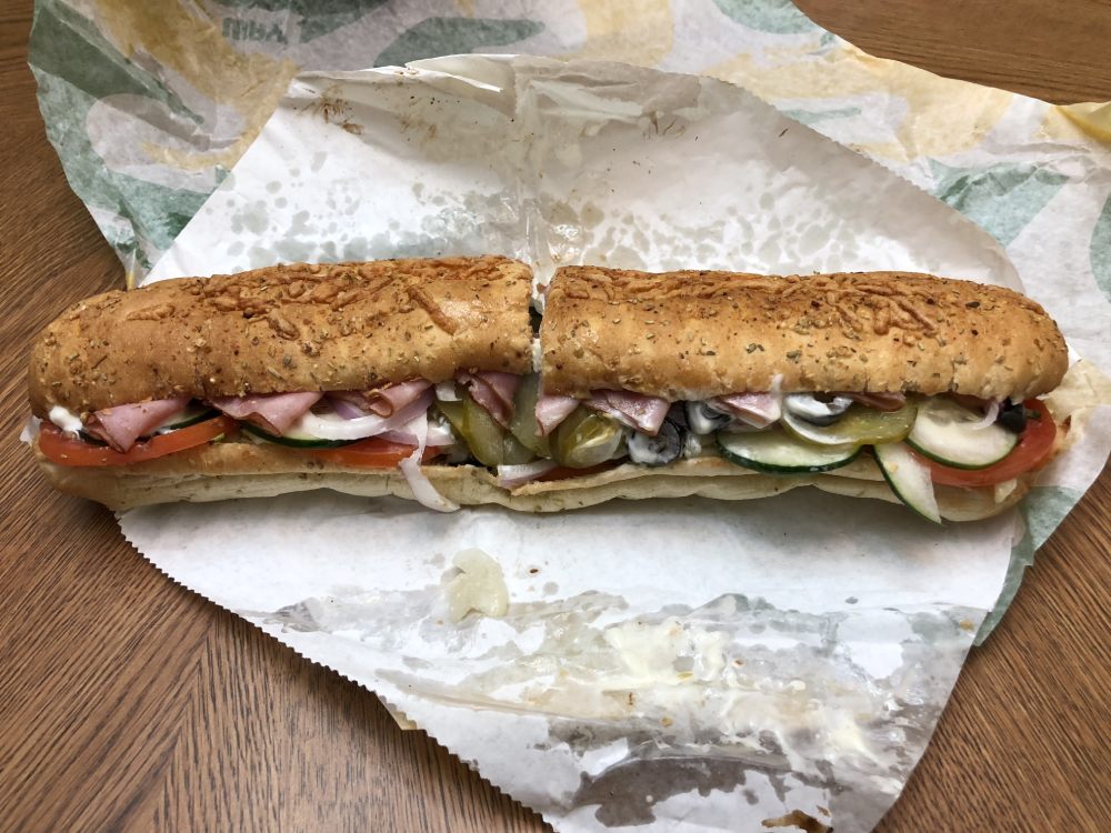 A cut-in-half Subway sandwich on a toasted, herb-topped roll filled with ham, tomatoes, cucumbers, pickles, and dressing.