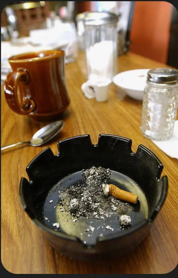 A black ashtray containing a cigarette butt and ash sits on a wooden table next to a brown coffee mug, a spoon, and a salt shaker.
