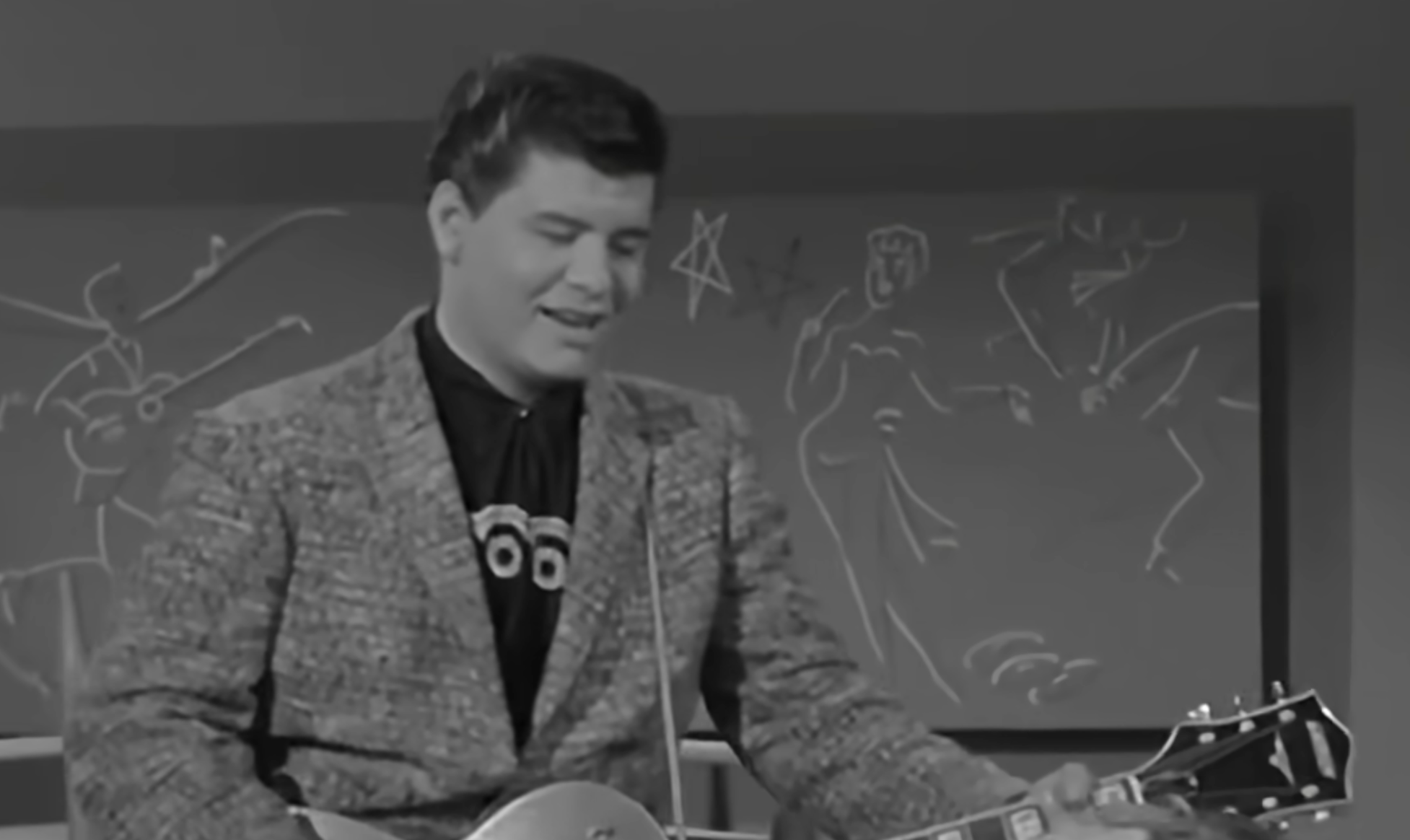 A man in a patterned blazer and a decorative shirt playing an electric guitar in front of a chalkboard with white line drawings.