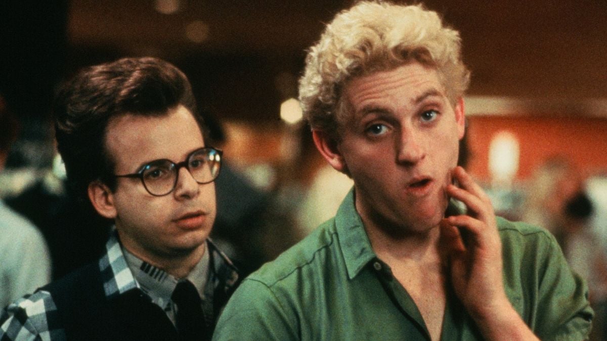 Two young men stand indoors; one wears glasses and a plaid shirt, looking serious, while the other, with blond curly hair and an open green shirt, looks surprised, touching his cheek. Background is blurry with people.
