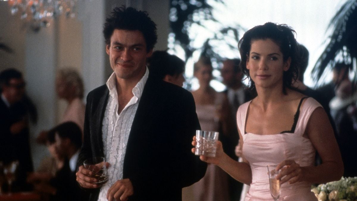 A man in a dark suit and a woman in a light pink dress hold drinks at a lively indoor party, with other guests chatting in the blurred background.