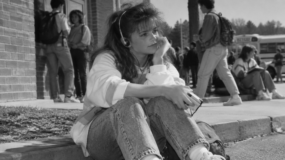 A girl with headphones sits on a curb, looking thoughtful while holding a portable cassette player, with other students and a school bus in the background.
