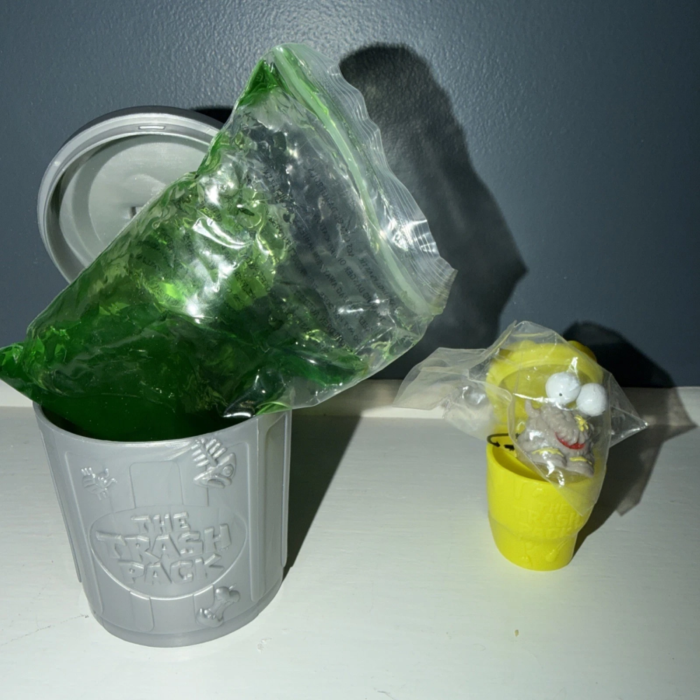 A grey Trash Pack toy trash can containing a bag of green slime, next to a small yellow container holding a toy figure in a plastic bag.