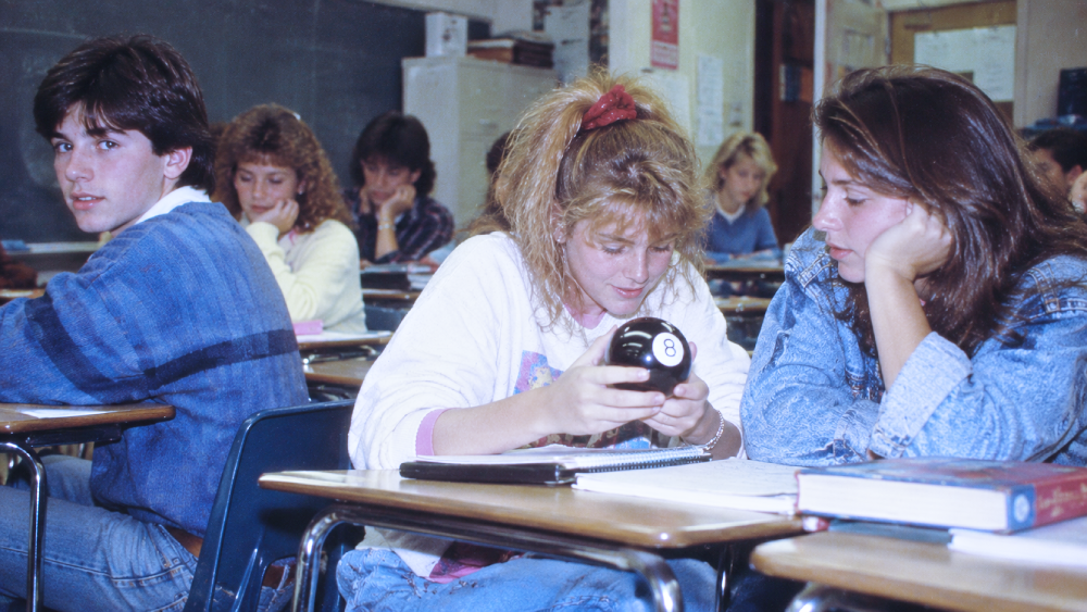 A classroom of students with a girl in the foreground looking at a Magic 8 Ball.