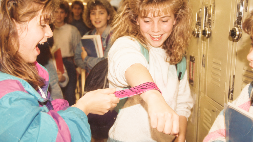 A girl places a pink patterned slap bracelet onto her friend’s wrist in a school hallway.