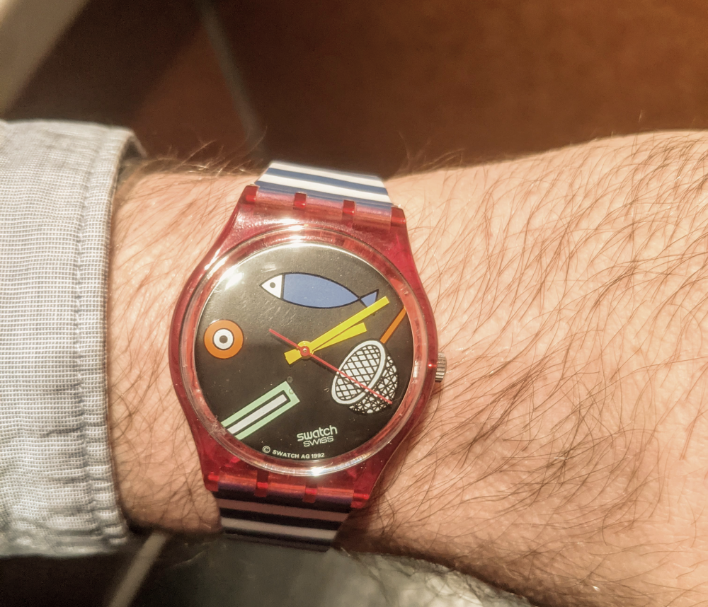 A Swatch wristwatch with a red translucent case and a black dial featuring a fish, a net, and abstract shapes, worn on a wrist with a striped strap.
