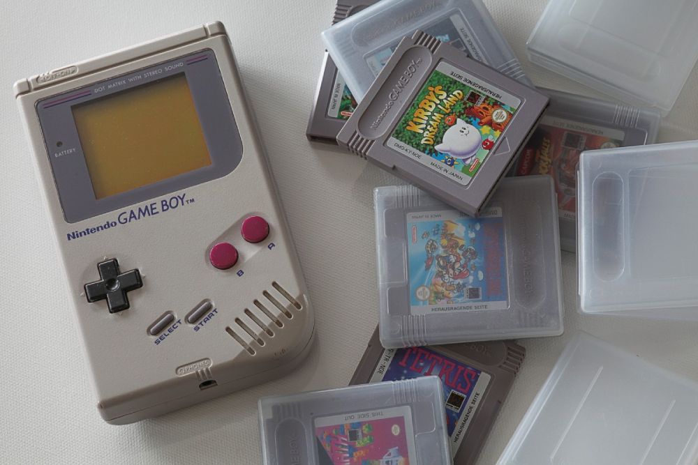 A grey original Nintendo Game Boy console surrounded by several Game Boy game cartridges and clear plastic storage cases.