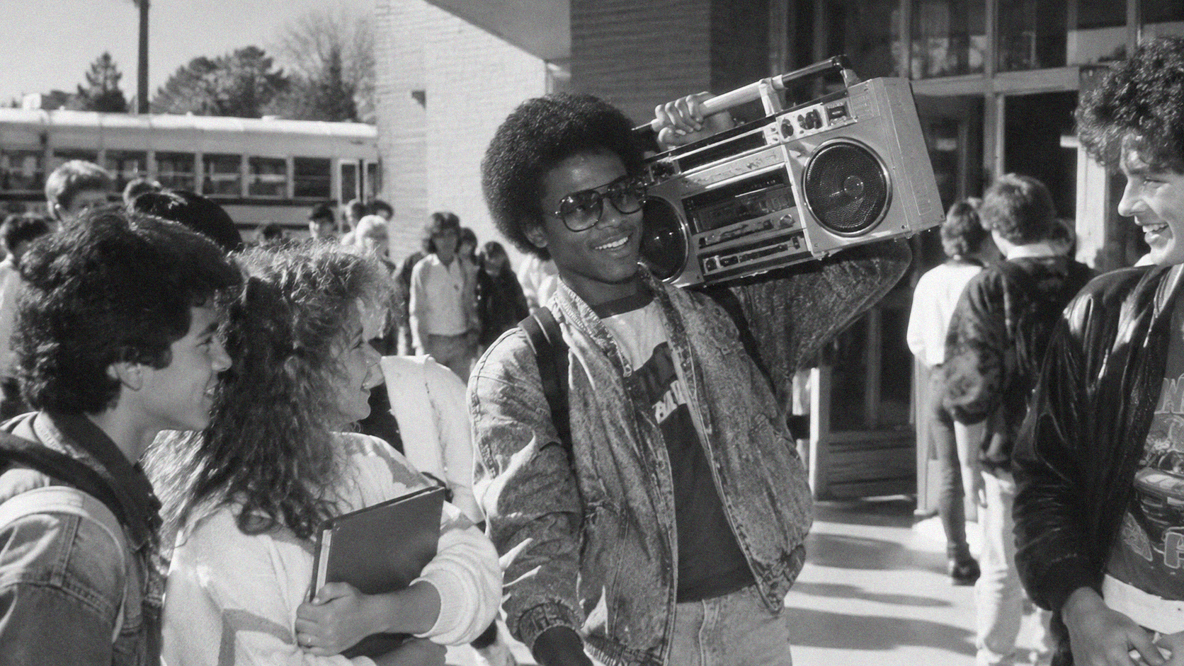 A smiling teenager with an afro and sunglasses carries a boombox on his shoulder while standing with friends outside a school.