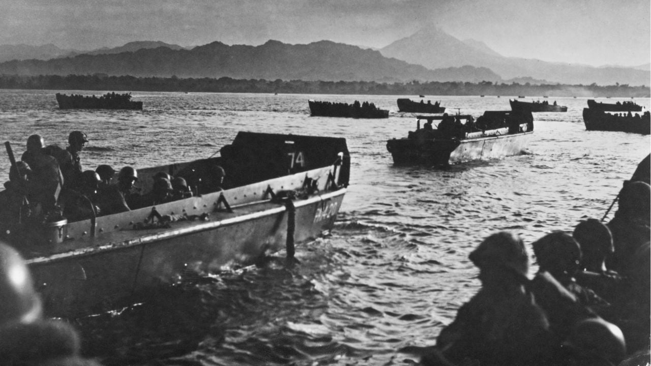 Soldiers in landing crafts approach a distant shore at dawn, with mountains in the background and several boats on the water, suggesting a military amphibious landing.