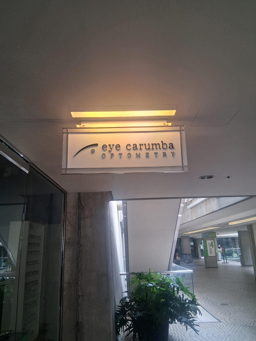 An illuminated sign for Eye Carumba Optometry hanging from the ceiling in a walkway.