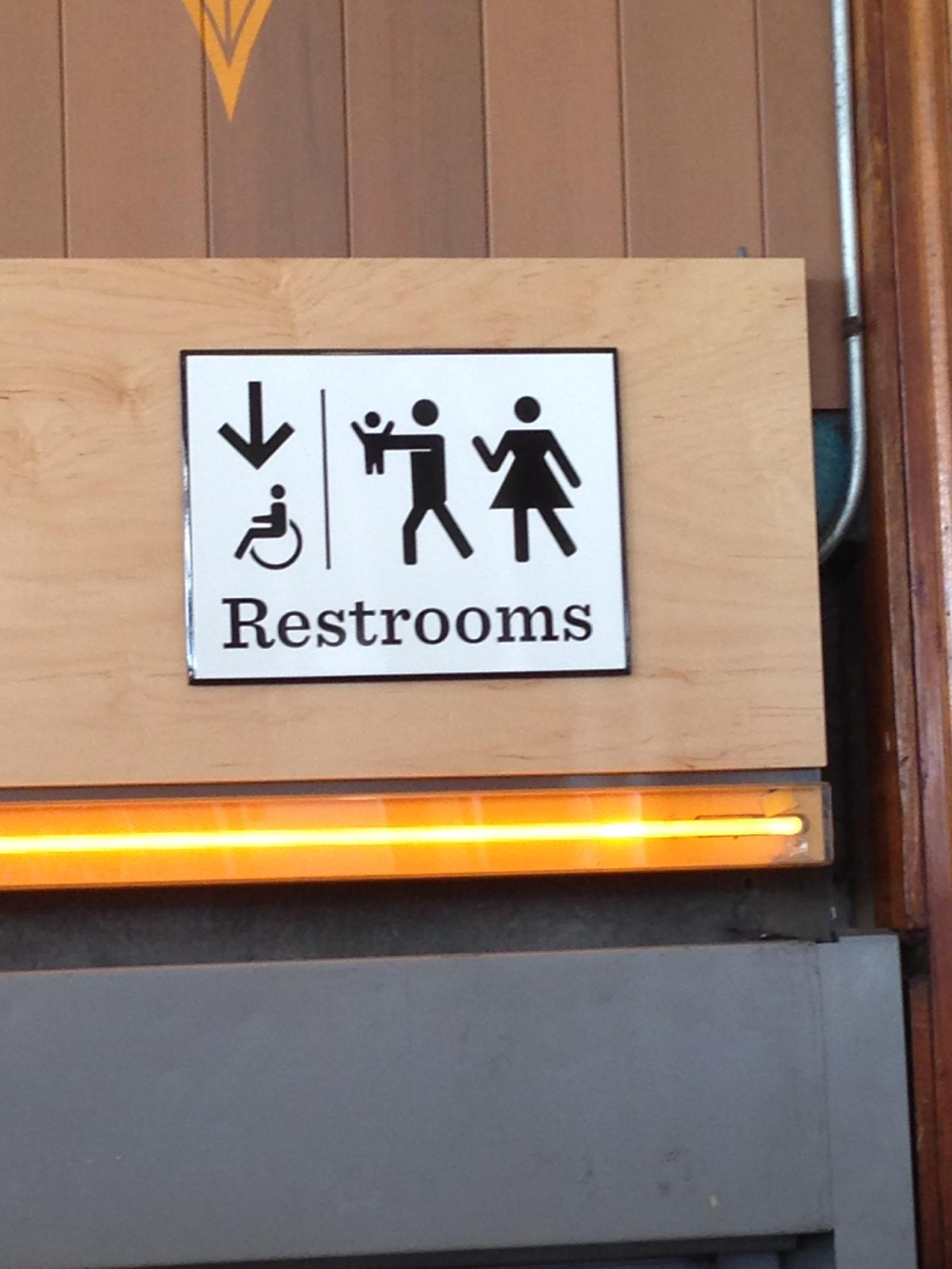 A restroom sign featuring icons for a wheelchair user, a person holding a baby, and a woman, with an arrow pointing downward.