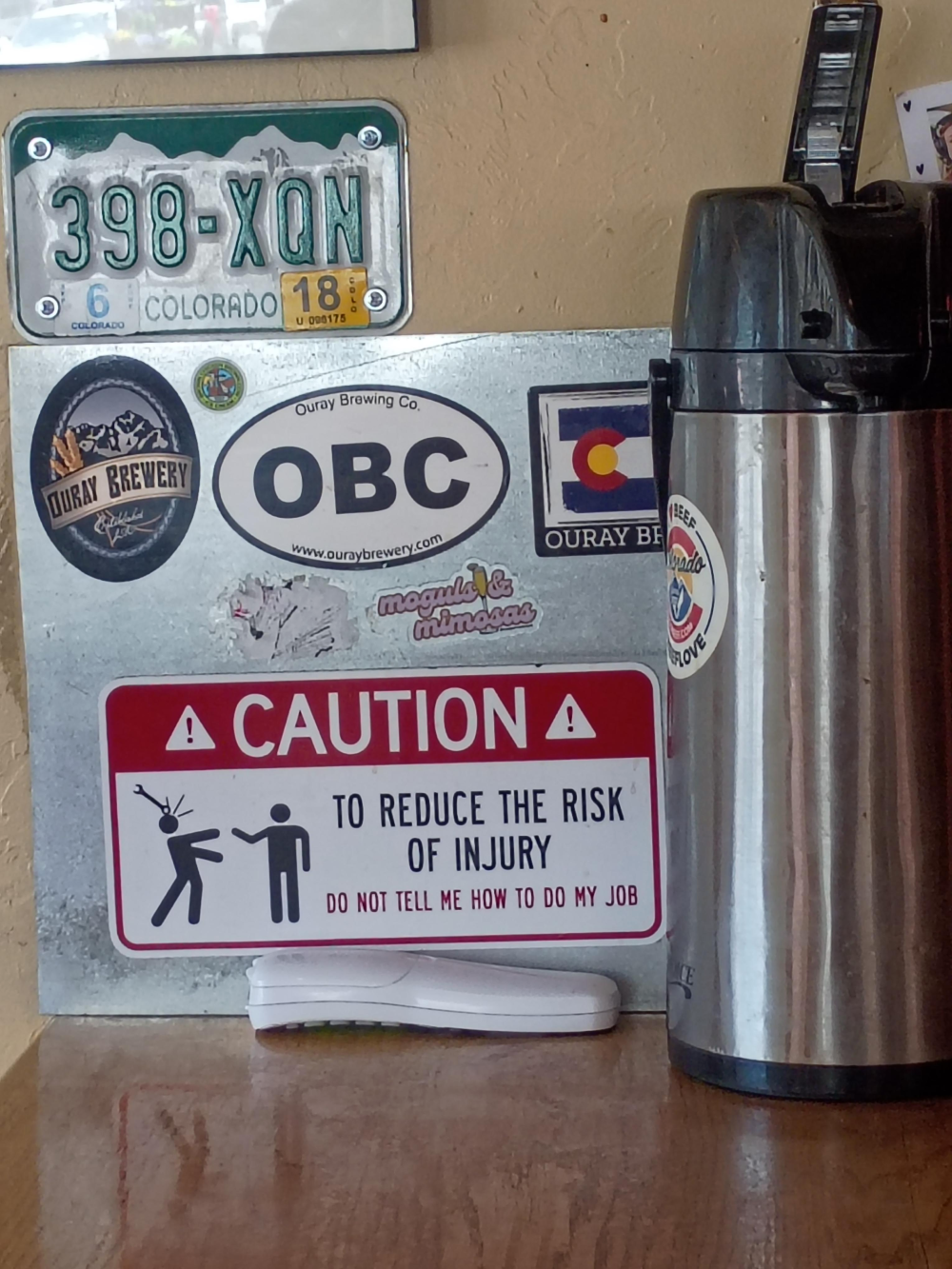 A metal sign covered in brewery stickers and a humorous warning label that reads, “Caution: To reduce the risk of injury, do not tell me how to do my job,” next to a metal coffee carafe and a Colorado license plate.