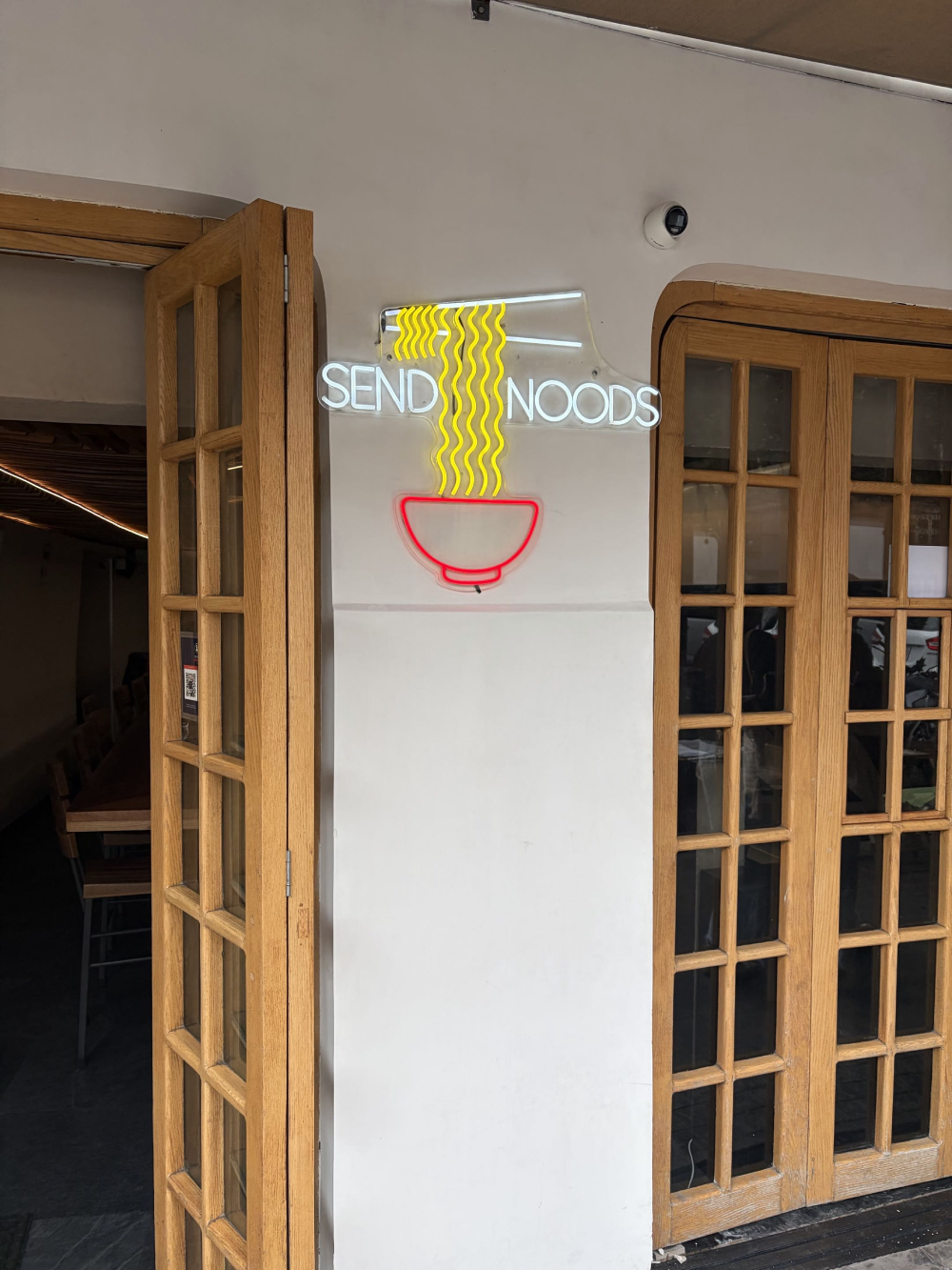 A neon sign reading “SEND NOODS” above a glowing red bowl of yellow noodles, mounted on a wall between two wooden-framed glass doors.