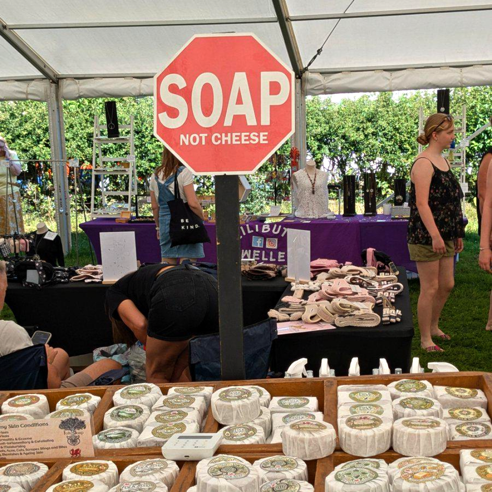 A red stop sign with the white text “SOAP NOT CHEESE” stands in front of a market stall selling wrapped soaps and accessories.