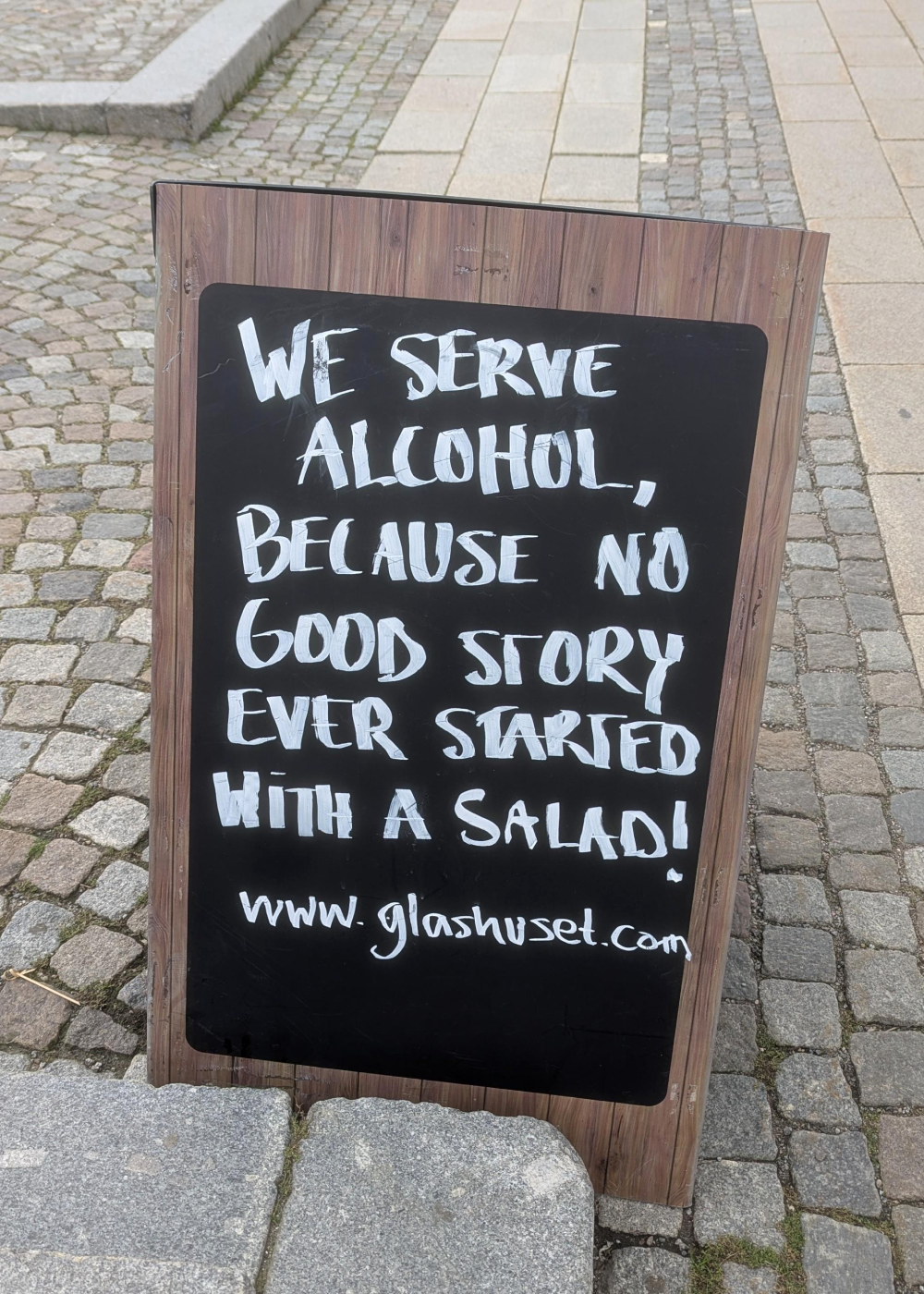 A sandwich board sign on a cobblestone path reading, “We serve alcohol, because no good story ever started with a salad!” followed by the website glashuset.com.