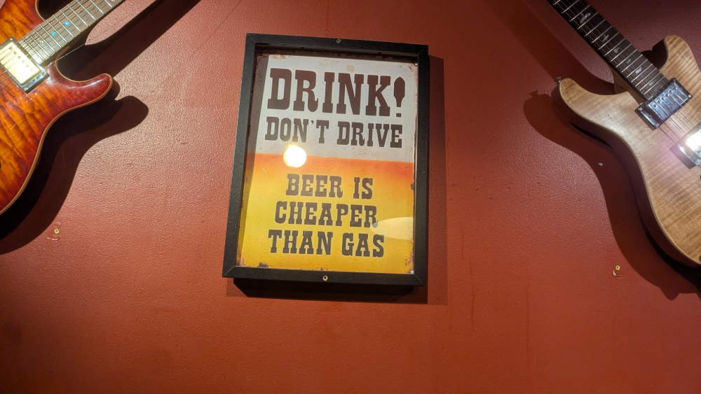 A framed sign on a red wall between two electric guitars that reads, “DRINK! DON’T DRIVE. BEER IS CHEAPER THAN GAS.”
