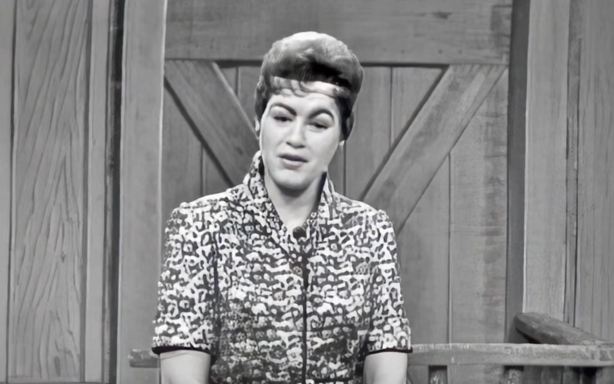 A woman with an upswept hairstyle wearing a patterned top sits in front of a wood-paneled wall.