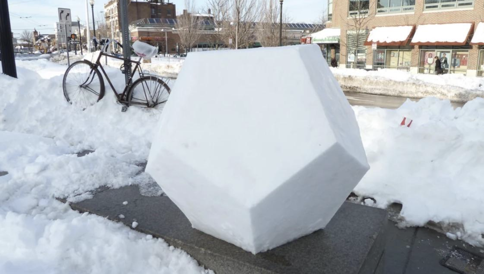 A dodecahedron carved from snow sitting on a stone pedestal in a snowy city street.