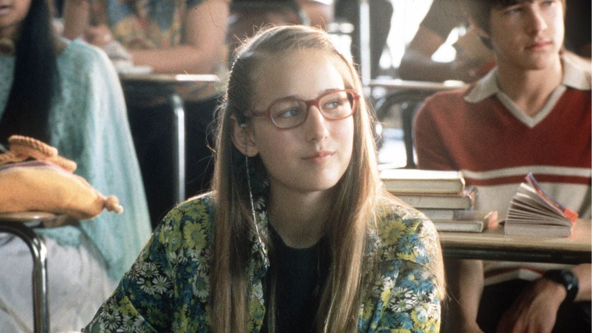 A teenage girl with long blonde hair and red glasses sits at a classroom desk, wearing a floral jacket. Other students and books are visible in the background.