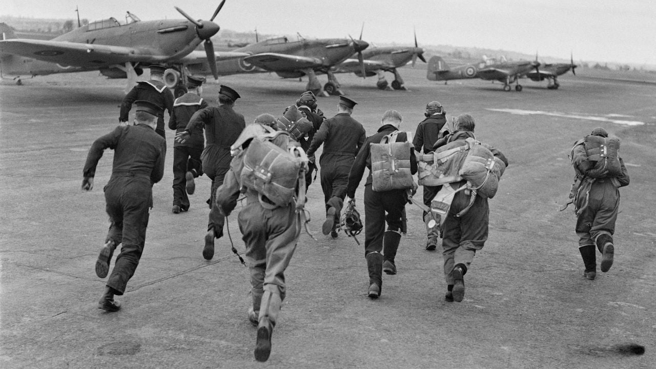 Pilots with parachutes run toward military aircraft lined up on an airfield, preparing for takeoff. The scene appears historical, with propeller planes visible and a cloudy sky overhead.