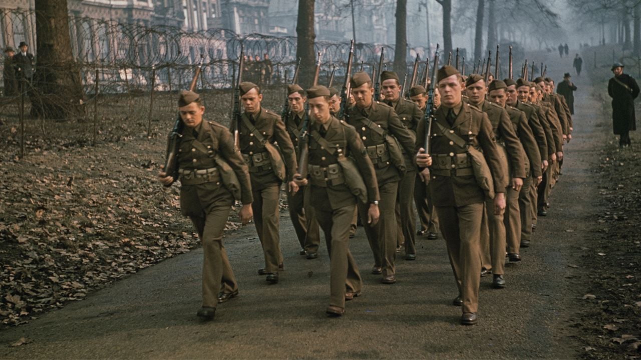 A group of soldiers in brown uniforms march in formation along a tree-lined path in a park, carrying rifles on their shoulders. The background shows barbed wire and buildings in a misty setting.