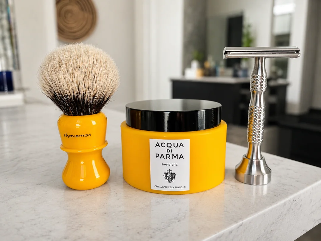 A yellow Shavemac shaving brush, a jar of Acqua di Parma shaving cream, and a metal safety razor arranged on a marble countertop.