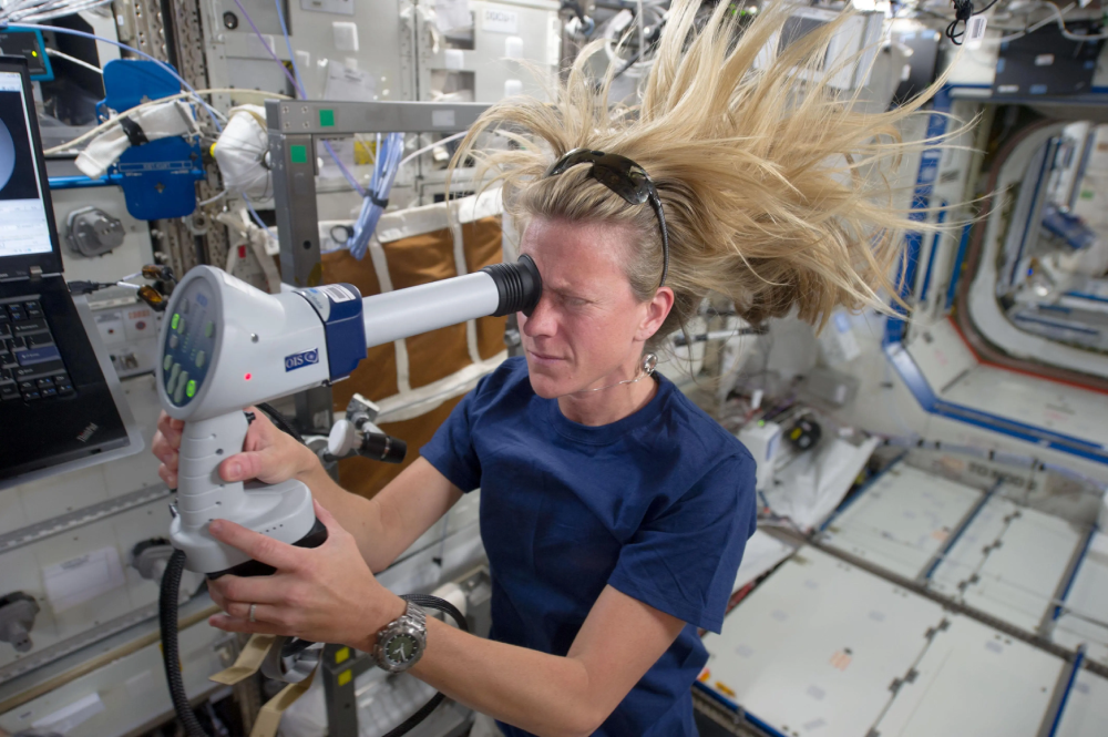 An astronaut with floating hair uses a handheld medical device to examine her eye while inside the International Space Station.