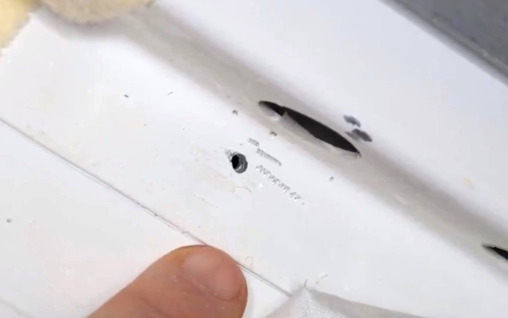 A finger points to a small hole in a white metal surface.